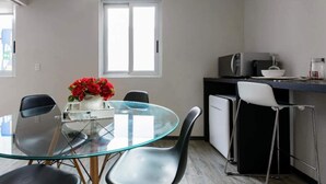 Un ambiente living compatto con un tavolo da pranzo in vetro, sedie nere, un angolo cottura dotato di forno a microonde e una finestra.