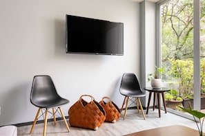Ein modernes Zimmer mit Flachbildfernseher an der Wand, zwei schwarzen Stühlen, einem kleinen runden Tisch mit einem Topf mit Pflanze und einem großen Fenster mit Blick auf Grünflächen.