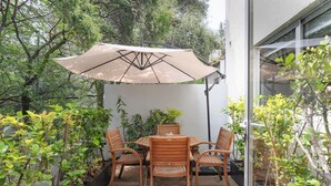 Um pátio com mesa e cadeiras, rodeado por plantas e um grande guarda-sol.