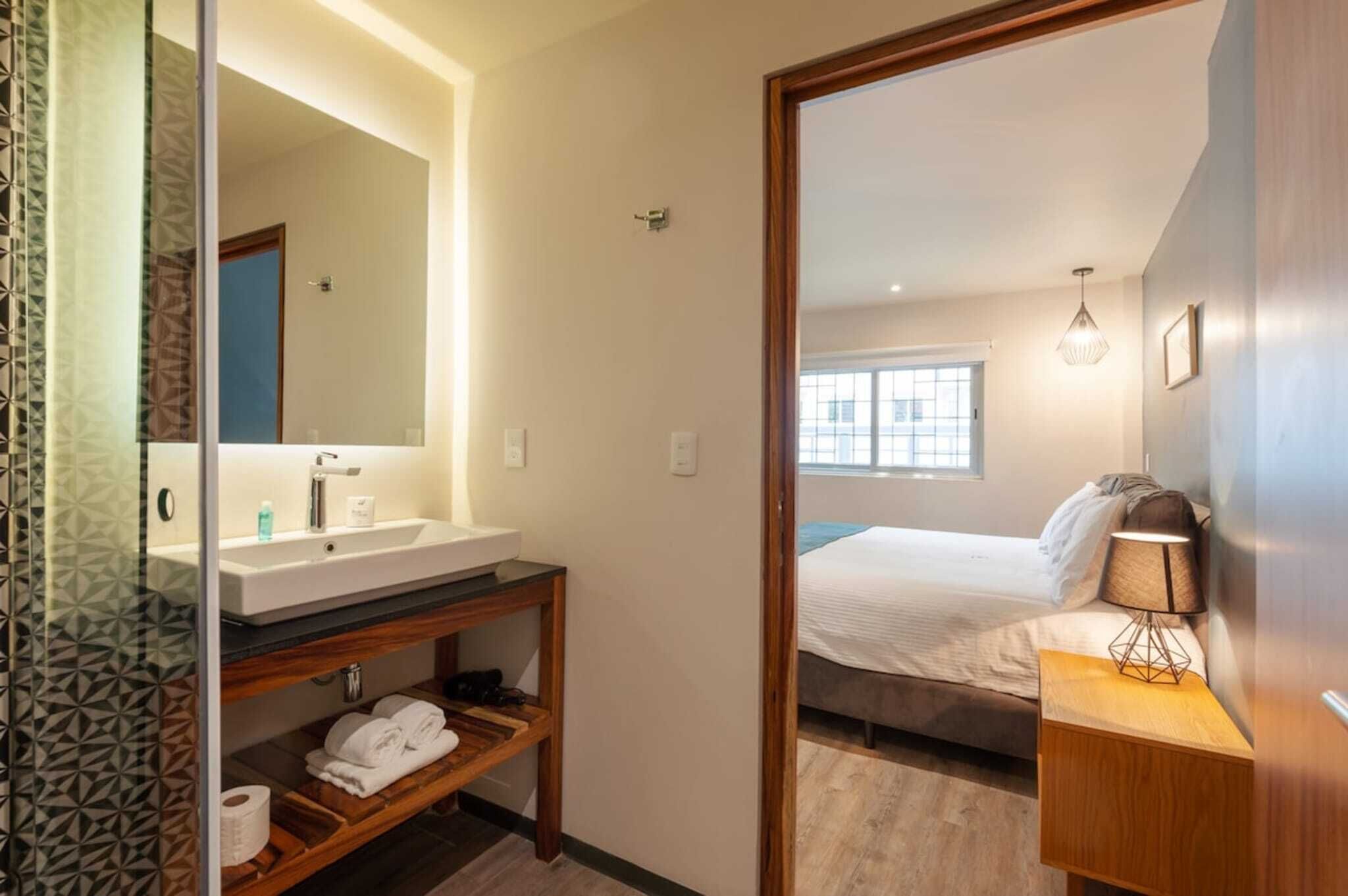 Quarto de hotel com banheiro, uma cama, mesa de cabeceira com abajur e uma janela.