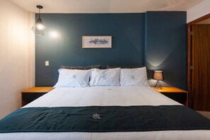Een net opgemaakt bed met een donker hoofdbord, een ingelijst beeld aan de muur en een bedlampje.