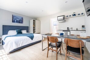 Ein modernes Hotelzimmer mit Bett, Schreibtisch, zwei Stühlen, einer Küchenzeile und einem Regal mit Geschirr.