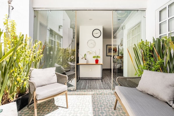A modern outdoor seating area with wicker furniture, potted plants, and a glass partition leading to an interior space with a clock and a small table.