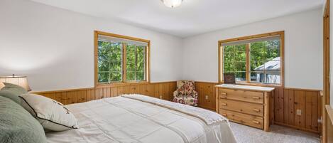 Un dormitorio con cama, dos ventanas, una silla, un tocador y una lámpara de techo.