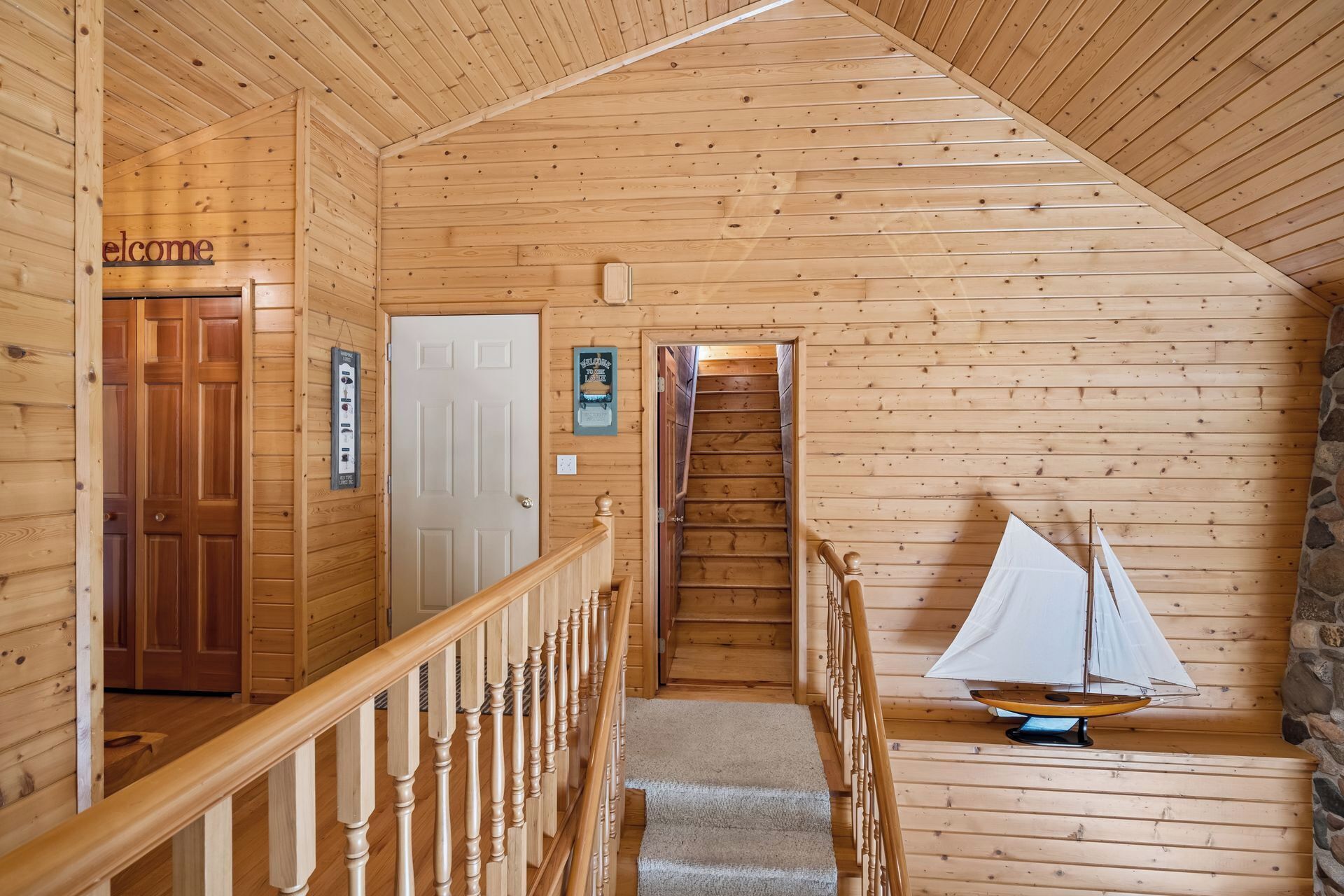 A wooden cabin interior with a staircase, a welcome sign, a door, and a sailboat model.
