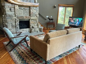 A living room with a stone fireplace, a sofa, two armchairs, a television, and a window with a view of trees. - Perfect house in peaceful Seeley Lake for a relaxing getaway (Seeley Lake)