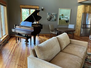A living room with a grand piano, a beige sofa, a dining table, and a refrigerator. - Perfect house in peaceful Seeley Lake for a relaxing getaway (Seeley Lake)
