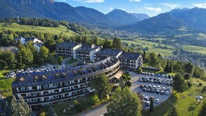 A hotel with a curved building, solar panels on the roof, a parking lot, and a mountainous backdrop. - Lucky Fox Loft - Residence Veronza (Cavalese)