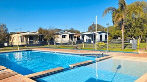 Una piscina con borde de azulejos, rodeada por un área cercada con edificios pequeños y árboles.
