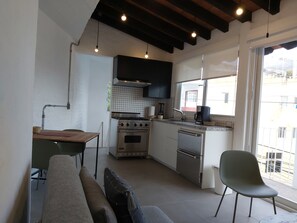 A compact kitchen with a gas stove, oven, and coffee maker, a dining table with chairs, and a view of the outside through a window. - Beautiful apartment with a view of the small town and just 10 minutes from the Romantic District. (Puerto Vallarta)
