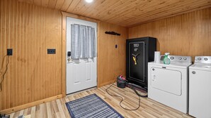 A compact room with wooden walls and ceiling, a white door with a curtain, a black safe, and a washing machine. - Doc's Place on Lark's:  Sleeps 4-Kayaks-Lakefront! (Pellston)