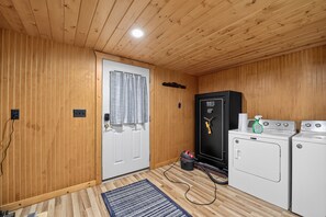 A compact room with wooden walls and ceiling, a white door with a curtain, a black safe, and a washing machine. - Doc's Place on Lark's:  Sleeps 4-Kayaks-Lakefront! (Pellston)
