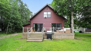 A red wooden house with a front deck, a grill, and a staircase leading to the door. - Doc's Place on Lark's:  Sleeps 4-Kayaks-Lakefront! (Pellston)