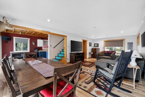 A spacious living area with a wooden dining table, a sofa, and a TV. - Doc's Place on Lark's: Tunnel of Trees Getaway | Lark's Lake | Lakefront | 2BR (Pellston)