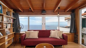 A cozy room with a red sofa, wooden bookshelves, a floor lamp, and a view of the sea. - Palheiro 360 – Stunning Bay View (Funchal)