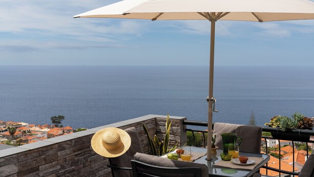 A balcony with a table, a chair, a large umbrella, and a view of the sea. - Palheiro 360 – Stunning Bay View (Funchal)