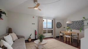 A living room with a sofa, a round table with chairs, a ceiling fan, a potted plant, and a view through a glass door. - Palheiro 360 – Stunning Bay View (Funchal)