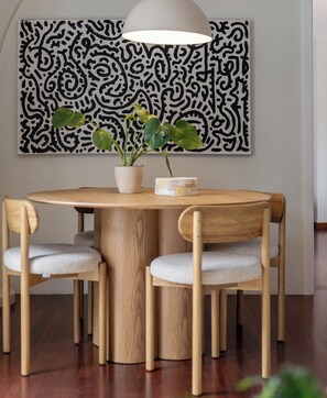 A dining area with a round wooden table, two chairs, a potted plant, and a wall-mounted abstract art piece. - Palheiro 360 – Stunning Bay View (Funchal)