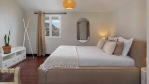 A bedroom with a bed, bedside table, potted plant, and a window with curtains. - Palheiro 360 – Stunning Bay View (Funchal)