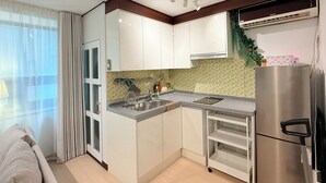 A compact kitchen with white cabinets, a stainless steel refrigerator, a built-in oven, and a window with curtains. - Modern Studio Near Gimpo Airport, King Bed + Pull-Out Trundle + Washer & Dryer (Seoul)