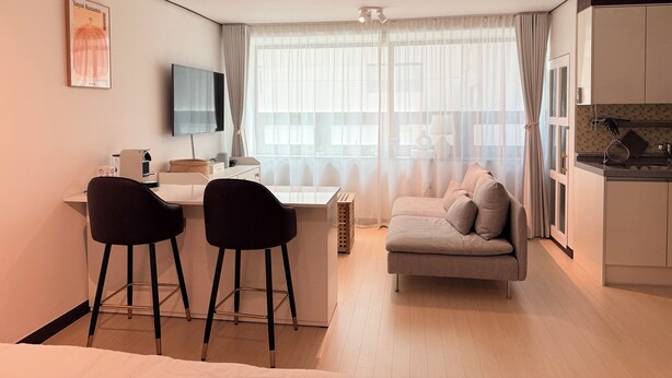 A modern hotel room with a desk, two chairs, a sofa, a TV, and a kitchen area. - Modern Studio Near Gimpo Airport, King Bed + Pull-Out Trundle + Washer & Dryer (Seoul)