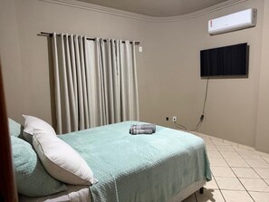 Quarto com cama, cortinas, ar-condicionado, televisão e tomada.