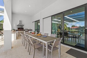 A modern outdoor dining area with a long table and chairs, a built-in grill, and a glass-enclosed patio with a view of a pool and palm trees. - Luxury Gulf Access Escape with Pool, Spa, Dock & Firepit Retreat - Villa Sogno - Roelens (Cape Coral)