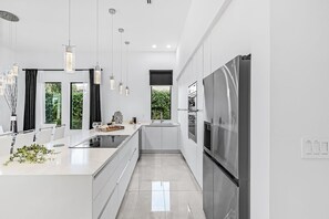 Une cuisine moderne dotée d’un îlot central, d’appareils électroménagers en acier inoxydable, de luminaires suspendus et offrant une vue sur la verdure environnante.