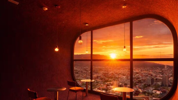 A room with a large window, round tables, and chairs, and a cityscape view at sunset.