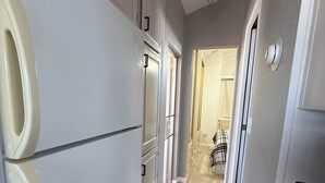 Un couloir étroit avec un réfrigérateur, une porte donnant sur une autre pièce et un luminaire au plafond.