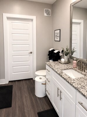 Un baño con puerta blanca, un inodoro, un lavabo, un espejo y una cita enmarcada en la pared.
