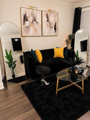 Ein modernes Wohnzimmer mit einer schwarzen Couch, gelben Kissen, einem Glastisch und zwei abstrakten Bildern an der Wand.