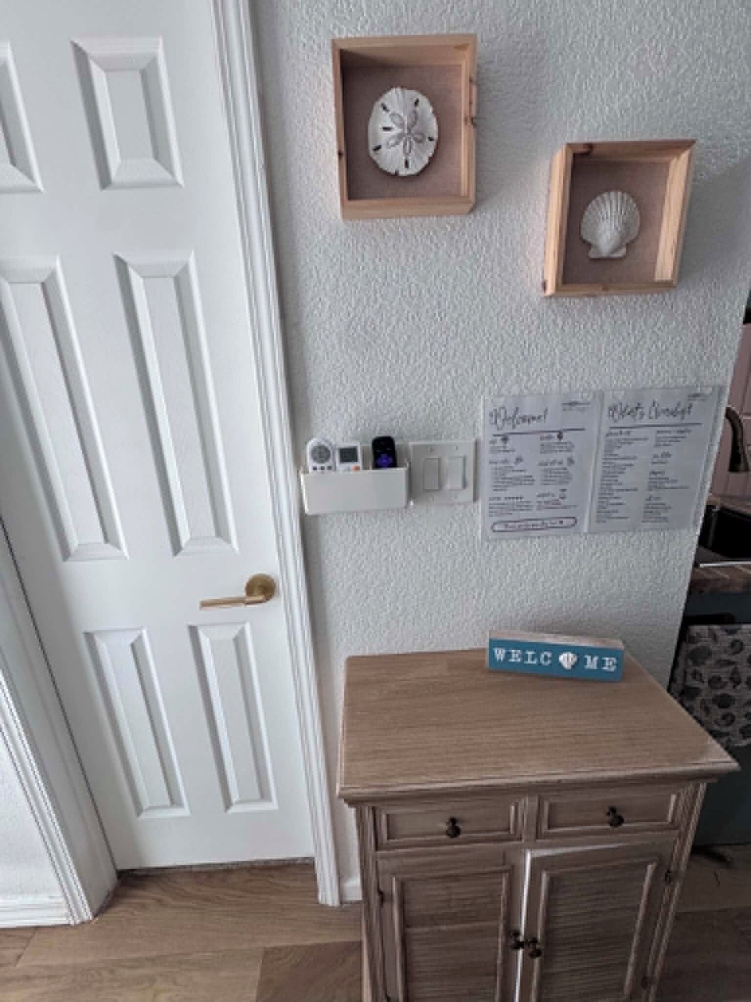 A room with a white door, a small wooden table with a 'WELCOME' sign, a wall-mounted clock, and a framed seashell.