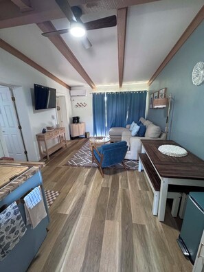 Una sala de estar con sofá, mesa de centro y televisor. Hay una mesa de comedor con un banco. El piso es de madera y tiene ventilador de techo.