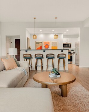 A modern living room with a round wooden coffee table, a sofa, and a kitchen area with a bar stool seating. - The King & Bear Retreat (St. Augustine)