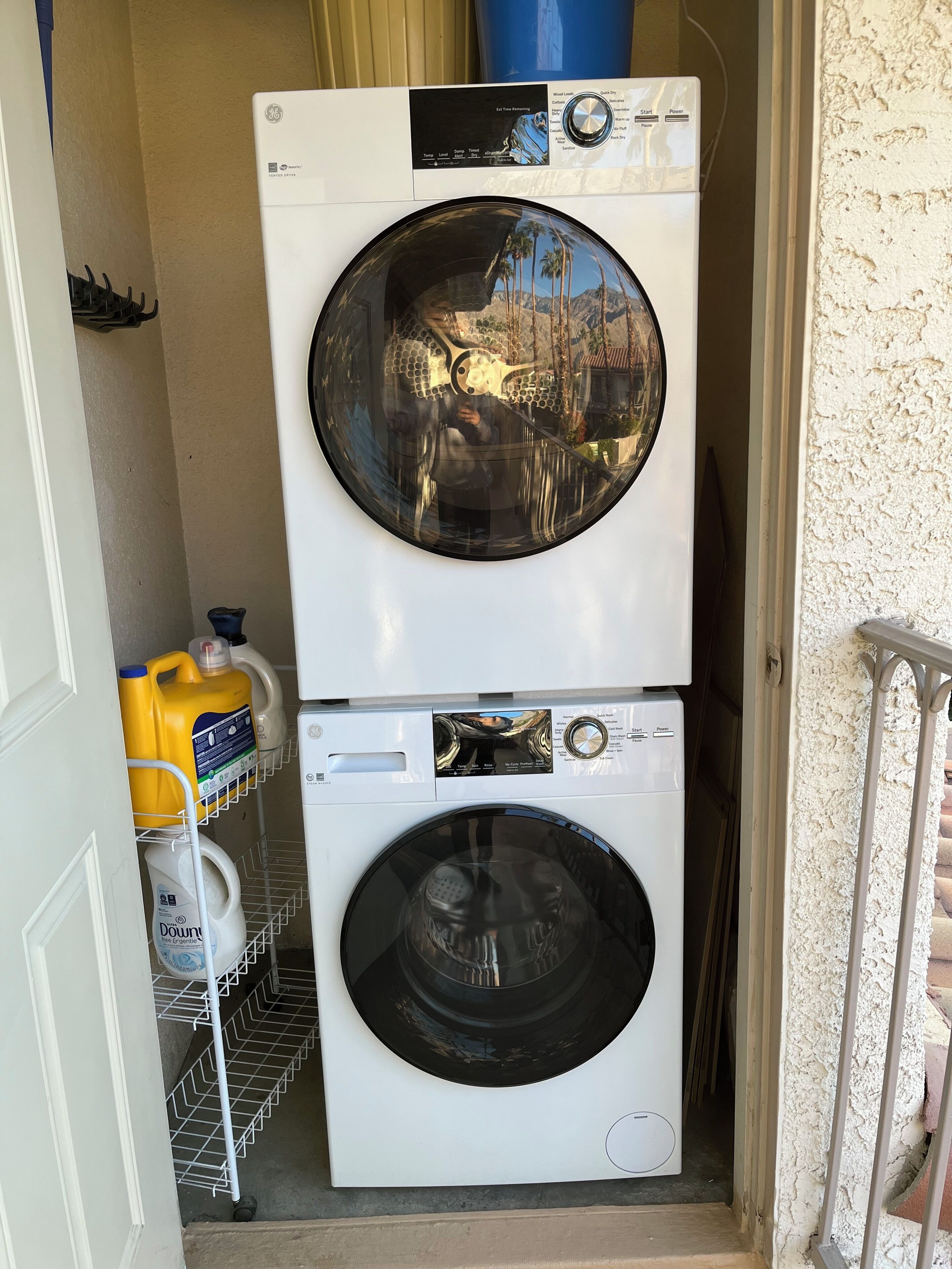 A stacked washer and dryer unit with a control panel, a metal shelf with cleaning supplies, and a metal railing.