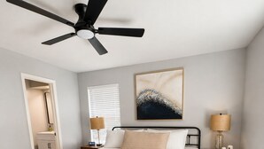 Ein Schlafzimmer mit einem Bett, Nachttischen, einem Deckenventilator, einem Gemälde und einer Tür zu einem Badezimmer.