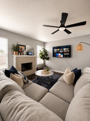 Ein modernes Wohnzimmer mit einer Eckcouch, einem Kamin, einem Deckenventilator und einem Wandfernseher, der verschiedene Kanäle zeigt.