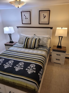 Una cama bien hecha con ropa de cama a rayas y almohadones decorativos, flanqueada por dos mesitas de noche con lámparas, y una obra de arte enmarcada en la pared de arriba.