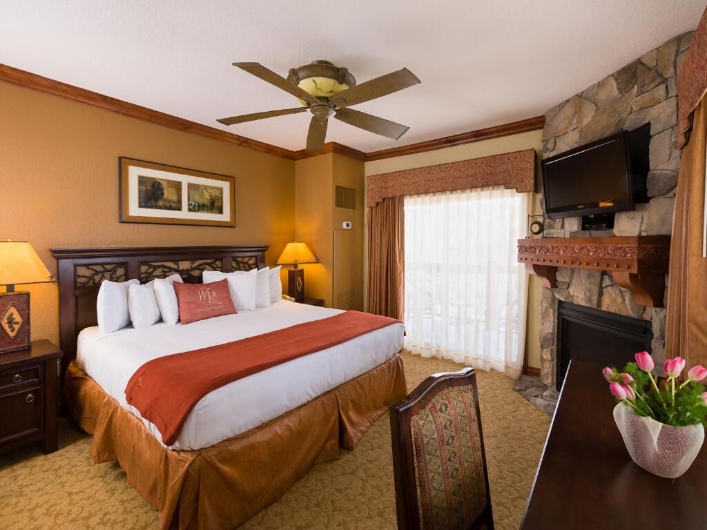 A hotel room with a bed, a ceiling fan, a fireplace, a television, and a vase with flowers.