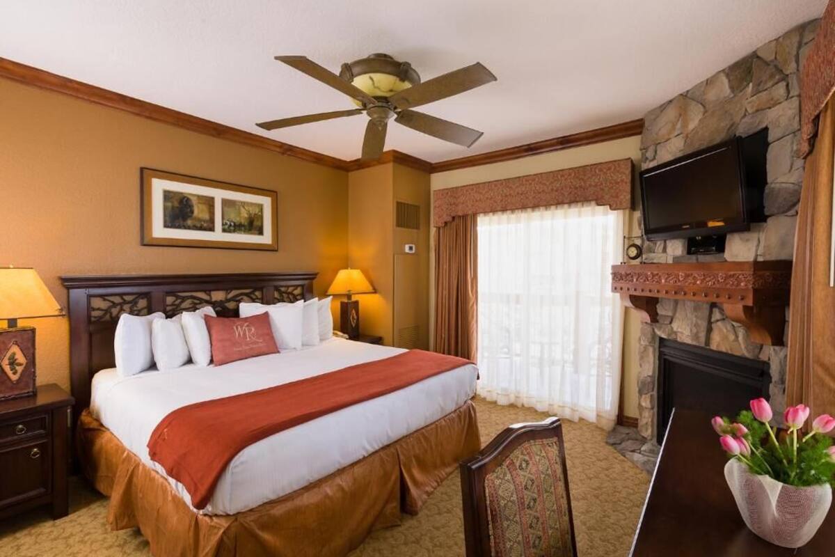 Ein Hotelzimmer mit einem Bett, einem Deckenventilator, einem Kamin, einem Fernseher und einer Vase mit Blumen.
