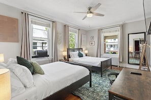 A hotel room with two beds, a desk, a chair, a ceiling fan, and large windows with curtains.