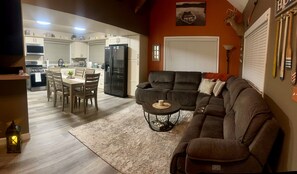 A living room with a sofa, a dining table with chairs, a kitchen area with appliances, and a fireplace.