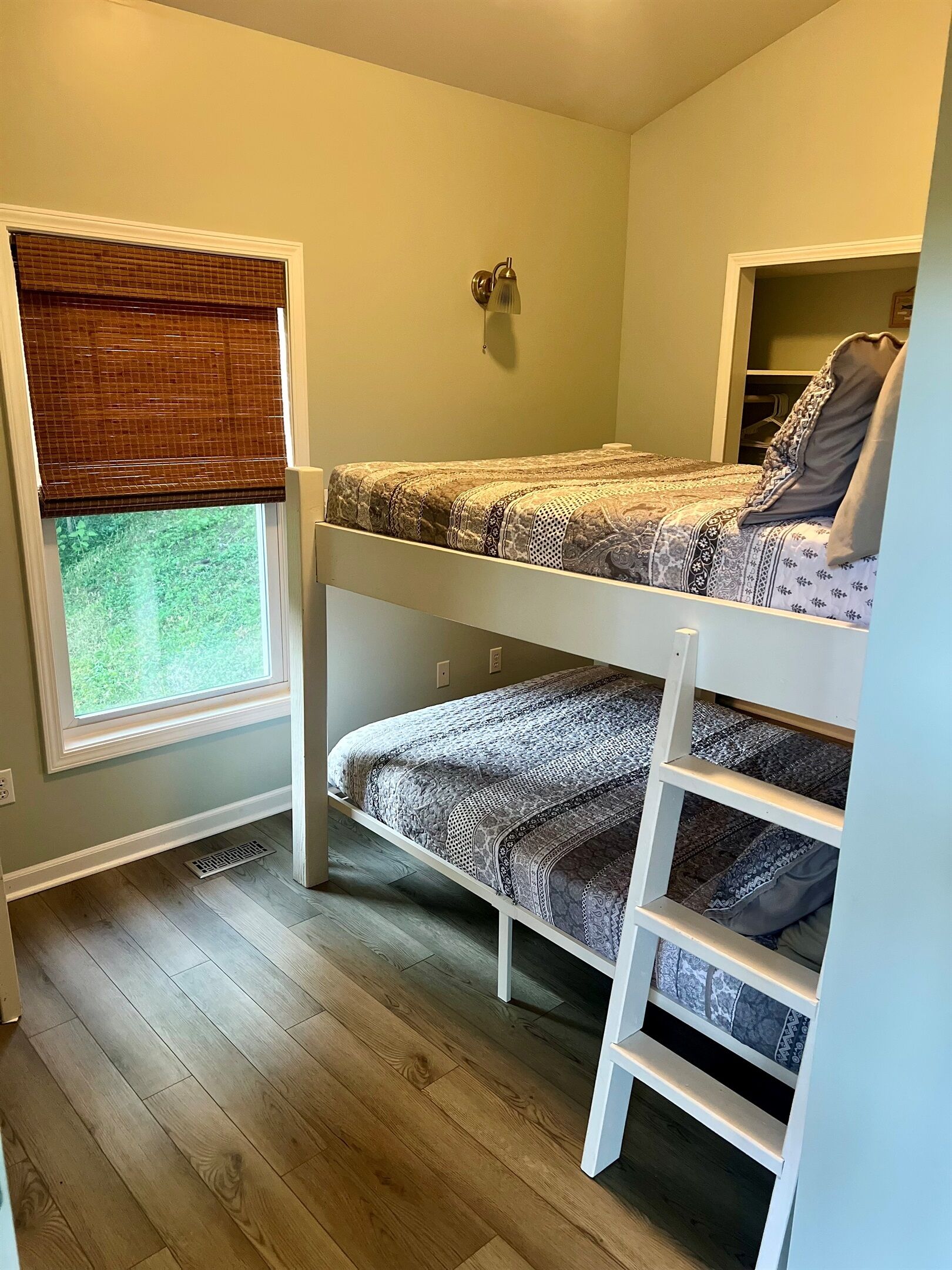 A room with a bunk bed, a window with a blind, a wall-mounted light, and a small shelf.