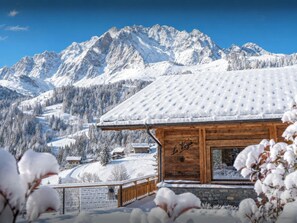 A wooden cabin with a snow-covered roof, surrounded by snow-covered mountains and trees.