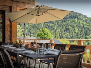 Un área de comedor al aire libre con una mesa preparada para cuatro, un parasol grande y vistas de una montaña boscosa.