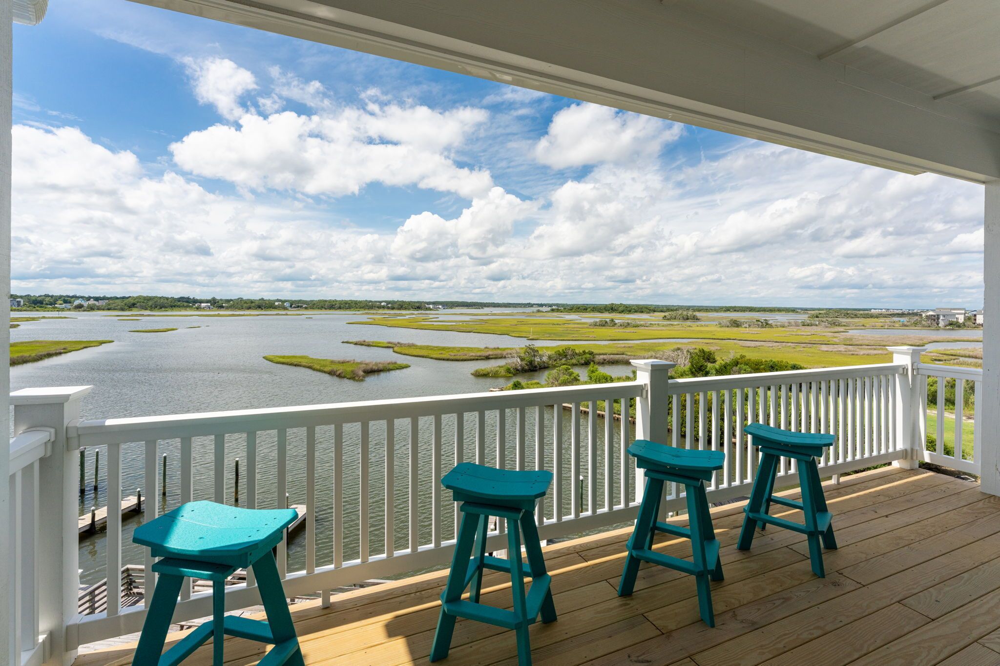Une terrasse en bois, dotée de rampes blanches et de trois tabourets turquoise, surplombe une étendue d’eau bordée de marais.