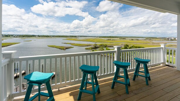A wooden deck with white railings and three turquoise stools overlooking a body of water with marshland. - Waterfront Paradise, 5BR, Elevator, Expansive Decks & Views (Surf City)