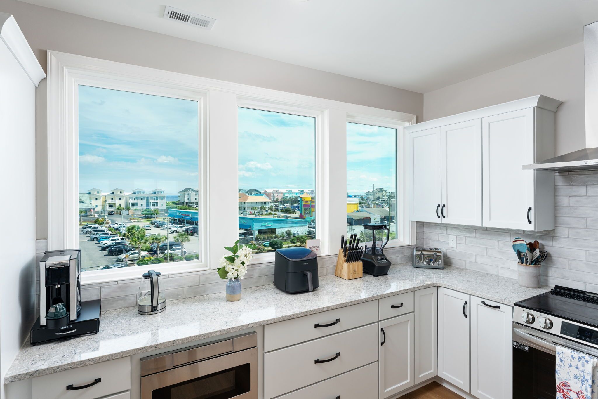 A kitchen with white cabinets, a stove, a coffee maker, and a view of a parking lot and buildings.