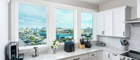 A kitchen with white cabinets, a stove, a coffee maker, and a view of a parking lot and buildings.